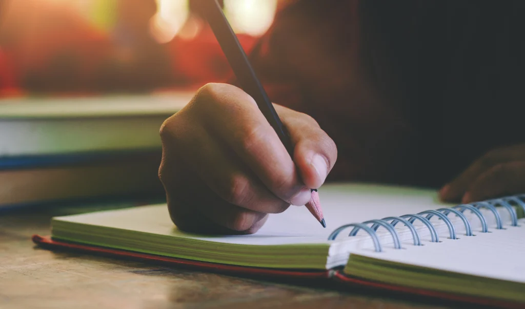person writing in a spiral notebook with a pencil