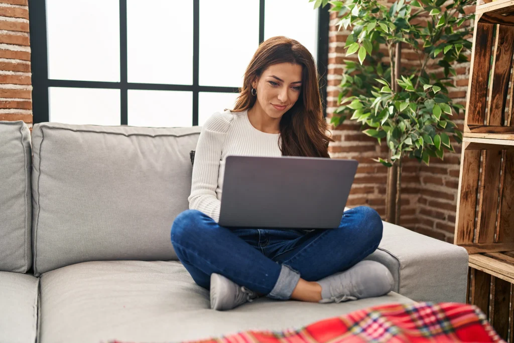 Woman smiling at her laptop. A bright, airy room with brick walls and a plant create a cozy setting.