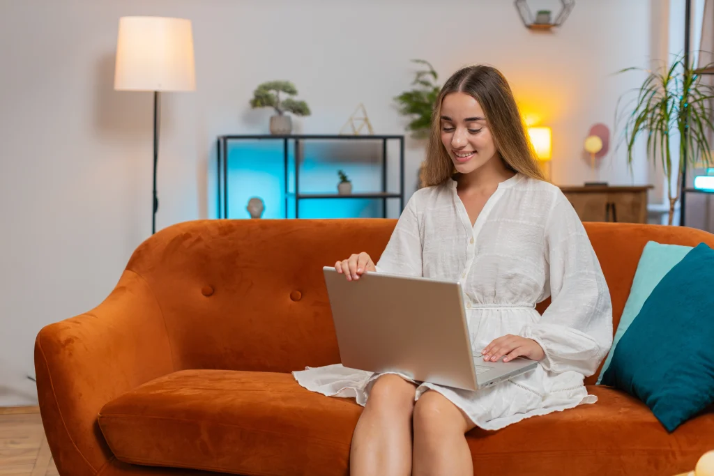 A woman using a laptop. The room is warmly lit with plants and shelves in the background.