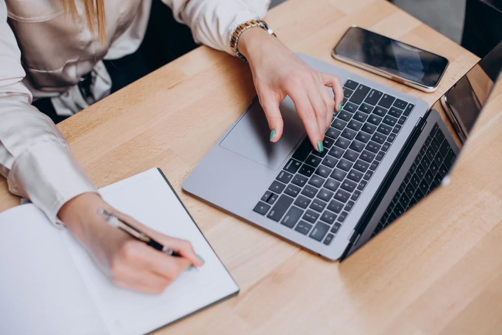 A person types on a laptop with one hand while writing in a notebook with the other; a smartphone is on the wooden desk.