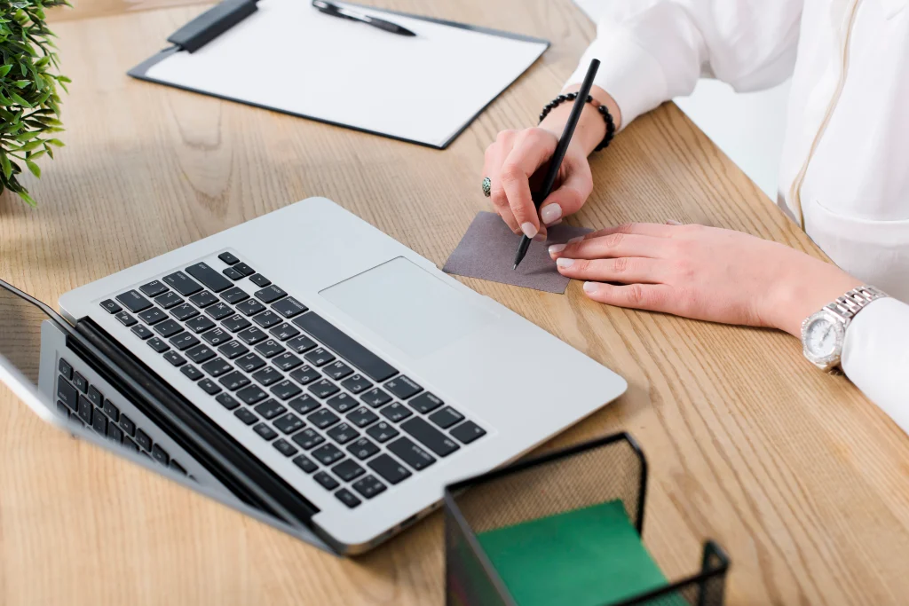 A person types on a laptop at a dark wooden table with a smartphone and a cup and saucer nearby.