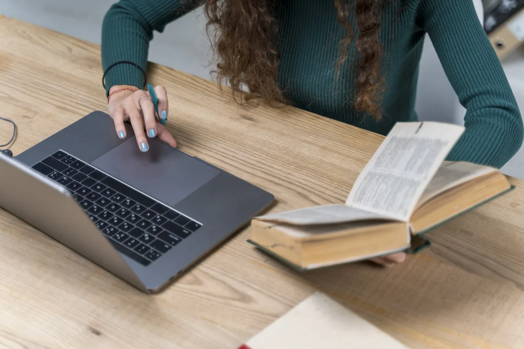 An overhead view of a woman at a desk, simultaneously typing on a laptop and referencing an open book.