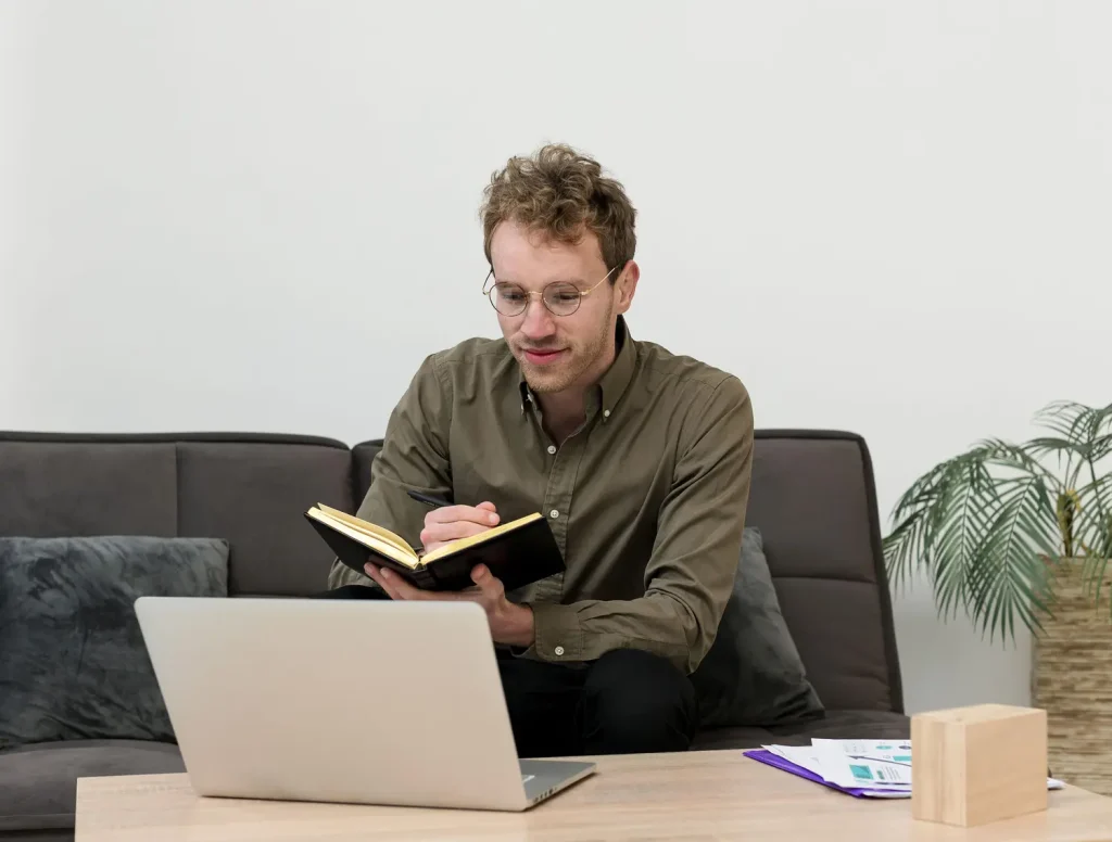 A man sits on a sofa with a laptop on the coffee table, holding a small notebook and looking thoughtful.