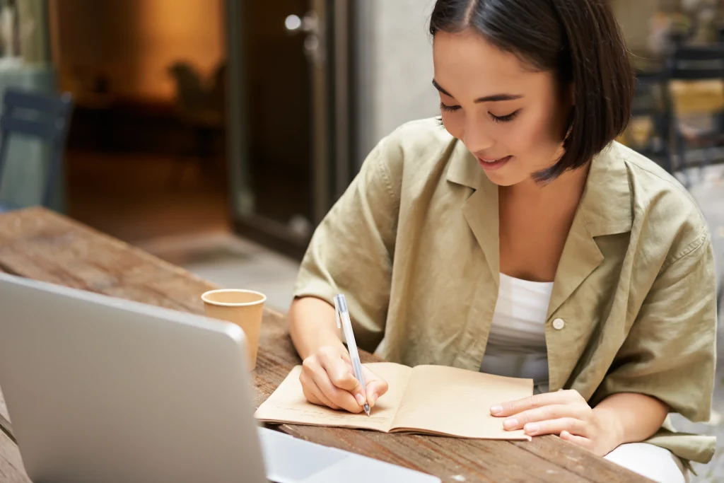 A woman in an olive shirt sits at a wooden table, writing in a notebook next to a laptop.